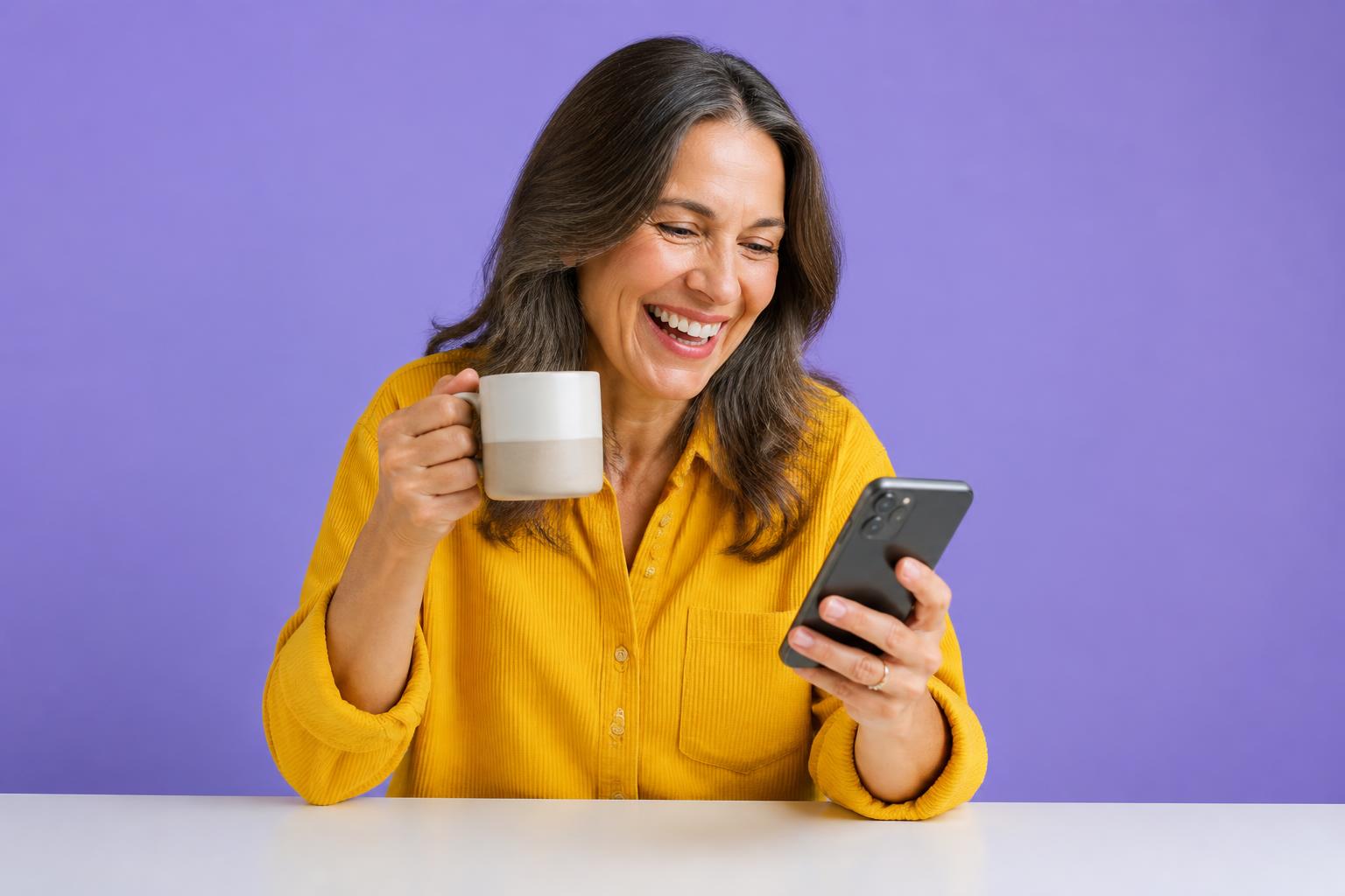 A woman in a yellow corduroy shirt holding a mug and laughing at her phone against a purple studio backdrop