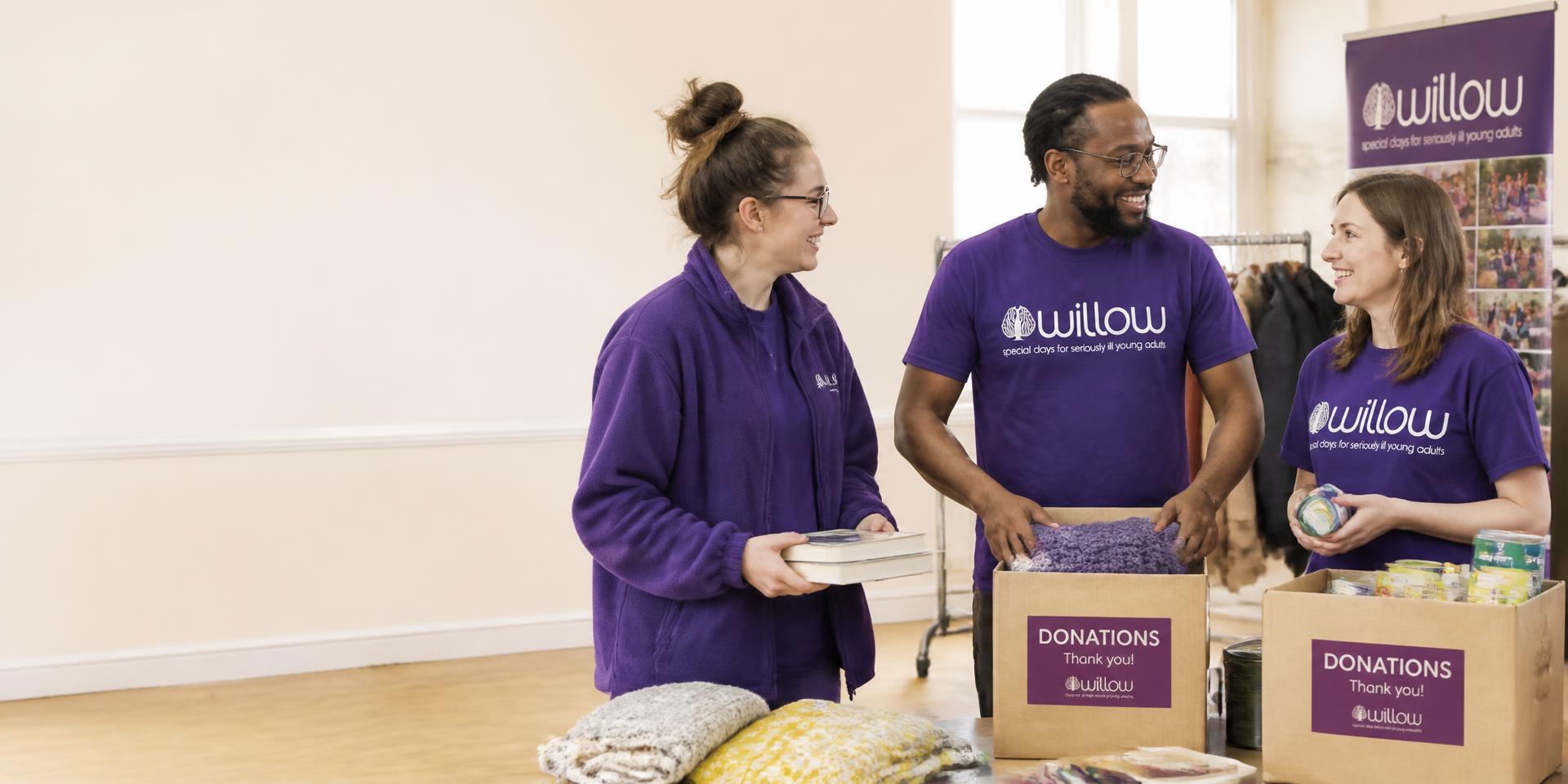 Three charity volunteers in purple branded tops sorting donation boxes in a bright community hall
